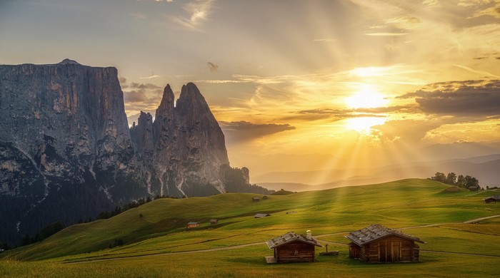 Wooden houses on grassy hills with mountains at sunset