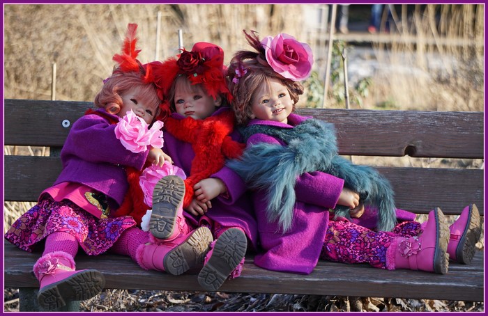 Three dolls dressed in purple and pink outfits sitting on a bench