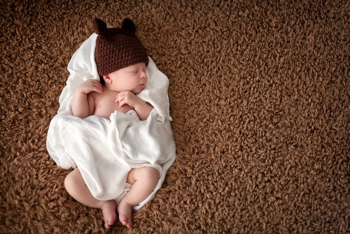 Newborn baby sleeping on a brown carpet wearing a hat