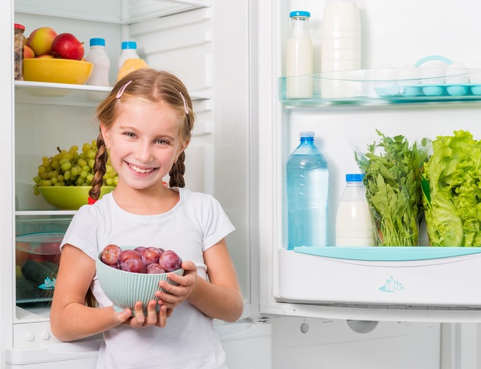 Girl holding plums in front of an open refrigerator
