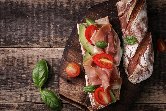Open-faced sandwich with ham, avocado, and tomatoes on a wooden board