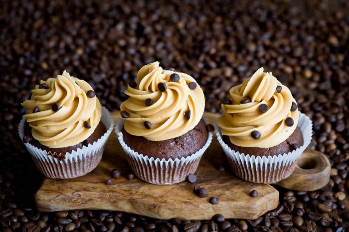 Three chocolate cupcakes with peanut butter frosting and chocolate chips on a cutting board surrounded by coffee beans
