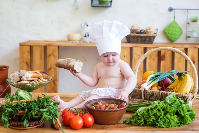 Baby in chef hat holding bread with fruits and vegetables