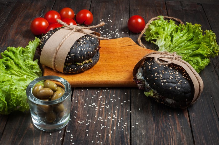 Two black hamburgers with sesame seeds on a wooden cutting board with tomatoes, lettuce, and olives