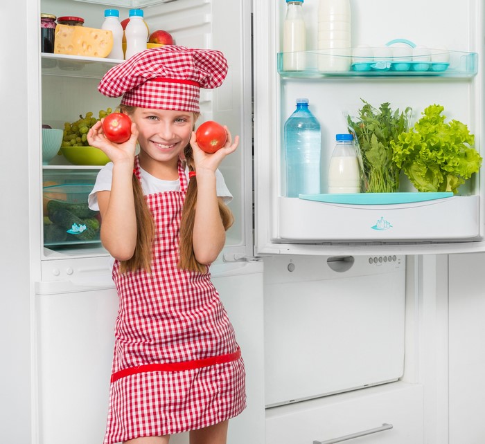 Girl in chef hat holding tomatoes in front of refrigerator