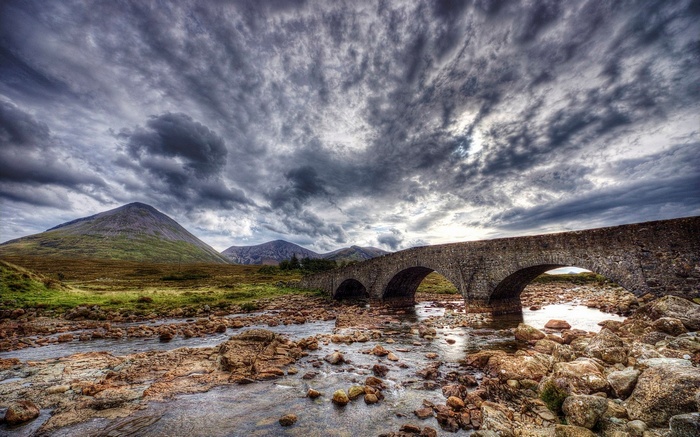 Stone bridge over a rocky river with mountains and stormy sky