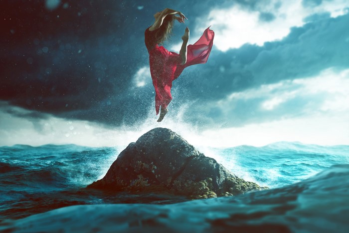 Woman in red dress jumps over a rock in the ocean