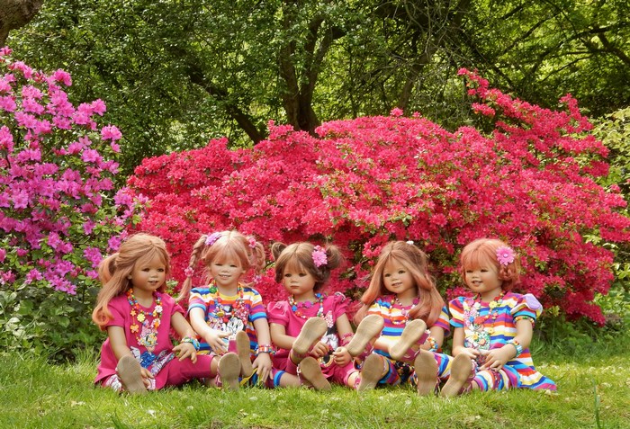 Five dolls sitting in front of pink rhododendrons