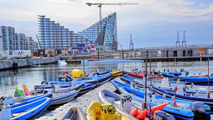 Boats docked at a marina with modern buildings and cranes in the background