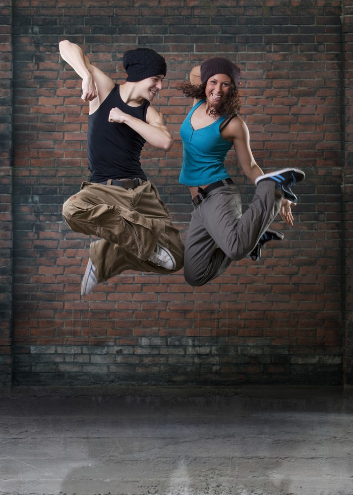 Two dancers in mid-air jump in front of a brick wall