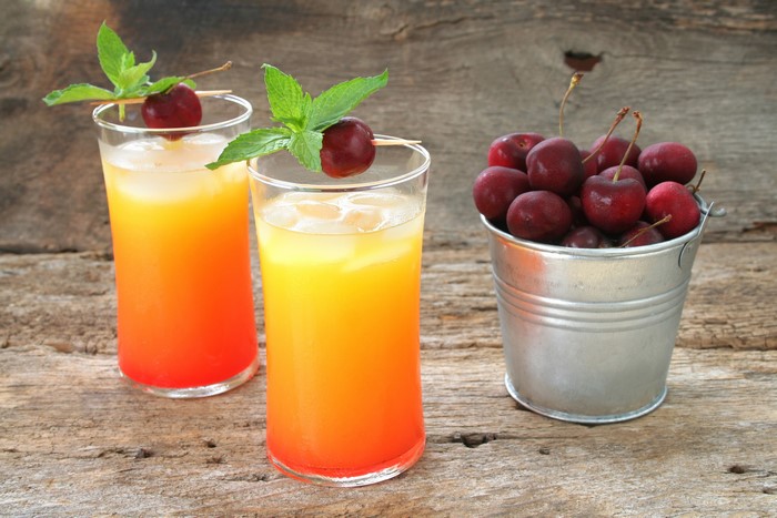 Two colorful cocktails with cherries and mint in glasses next to a bucket of cherries on a wooden table