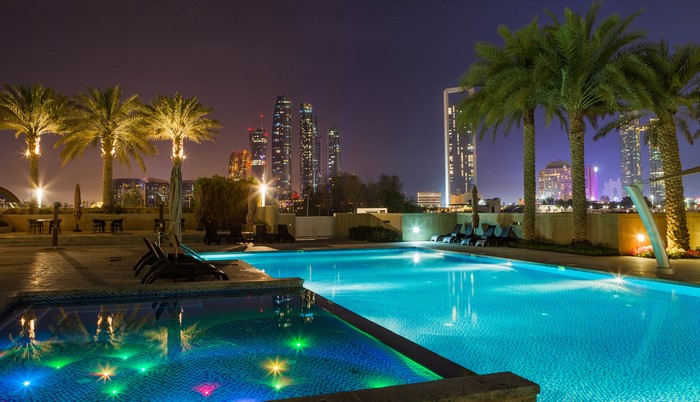 Resort pool with palm trees and city skyline at night in Abu Dhabi