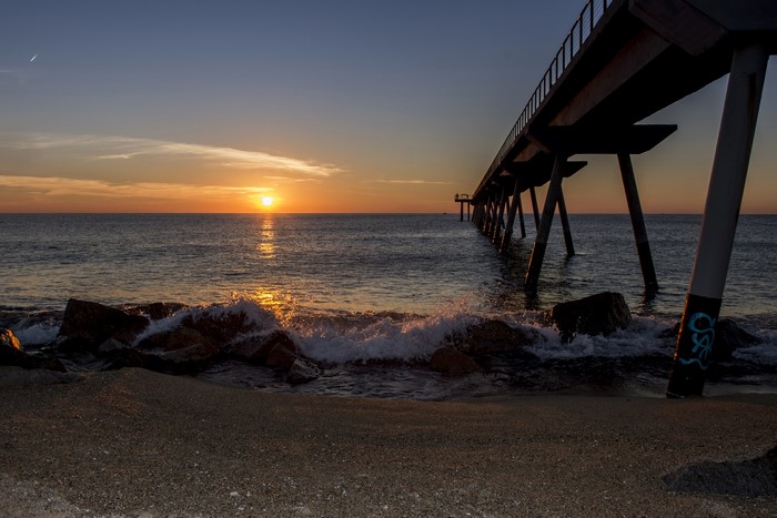 Sunrise over the sea with a pier and waves crashing on rocks