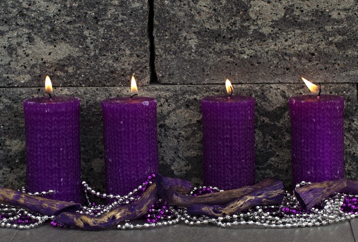 Four purple advent candles lit in front of a stone wall