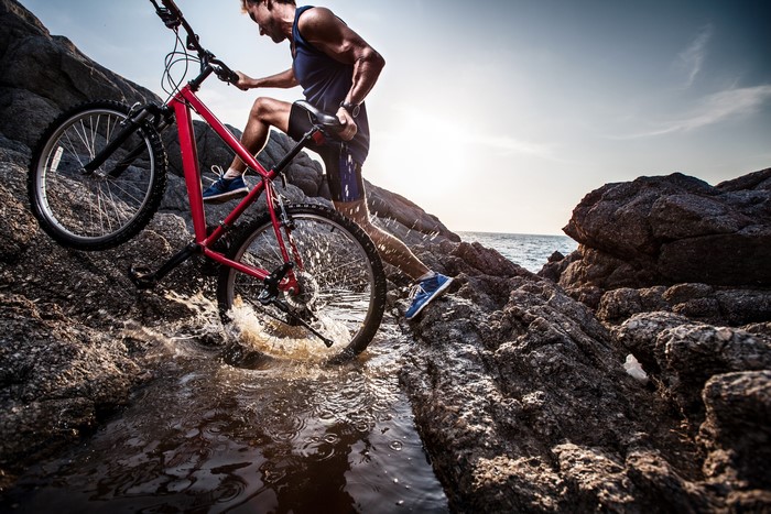 Man carrying bicycle through rocky terrain near water
