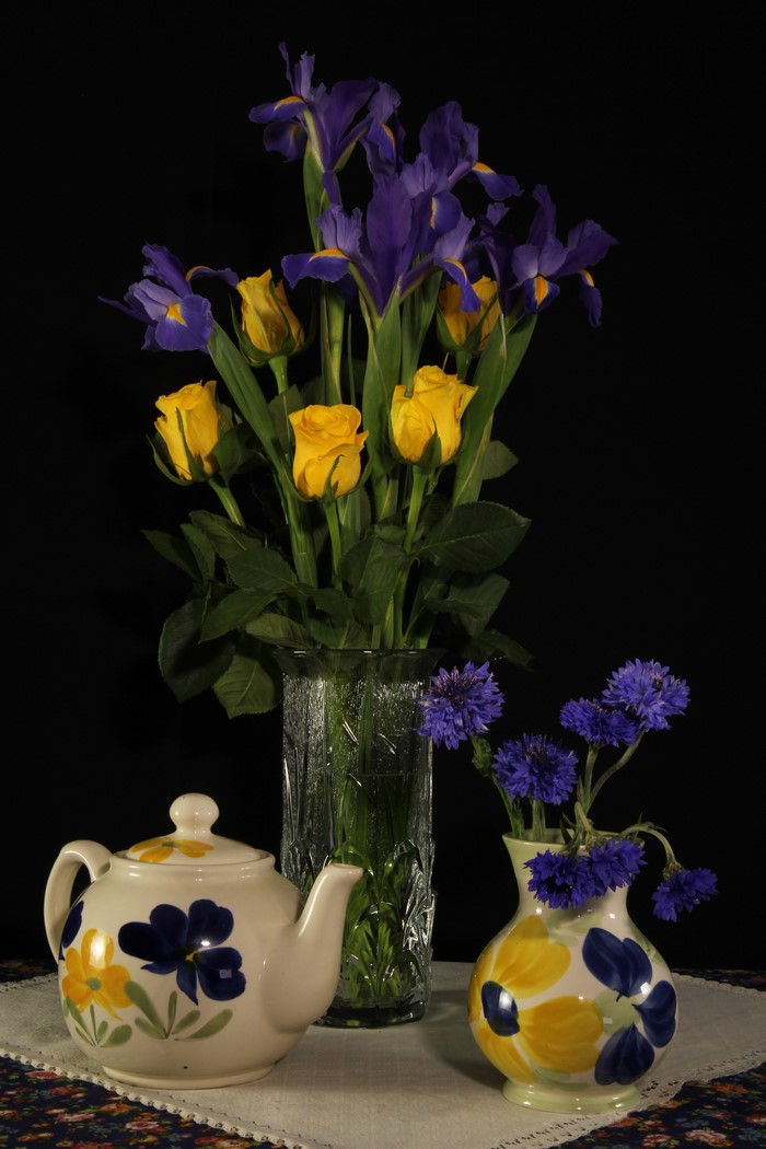 Still-life with yellow roses, purple irises, and cornflowers in vases with a teapot