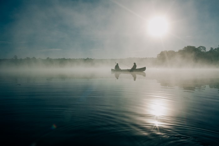 Two people fishing in a canoe on a foggy lake at sunrise