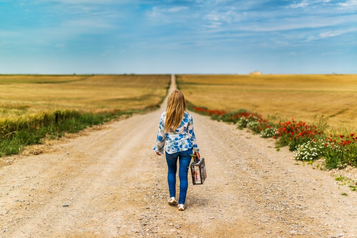 Girl with suitcase walking on a dirt road through fields
