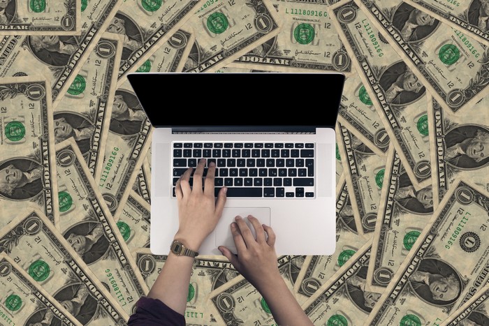 Hands typing on a laptop surrounded by dollar bills