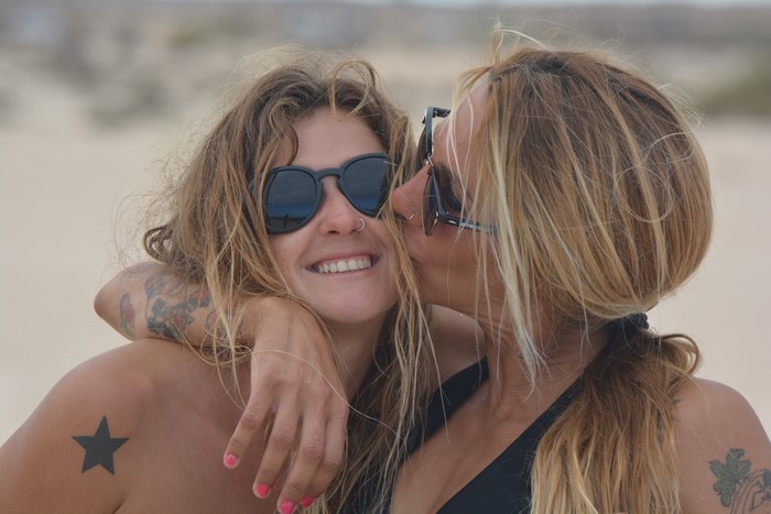 Two women with tattoos and sunglasses hugging on a beach