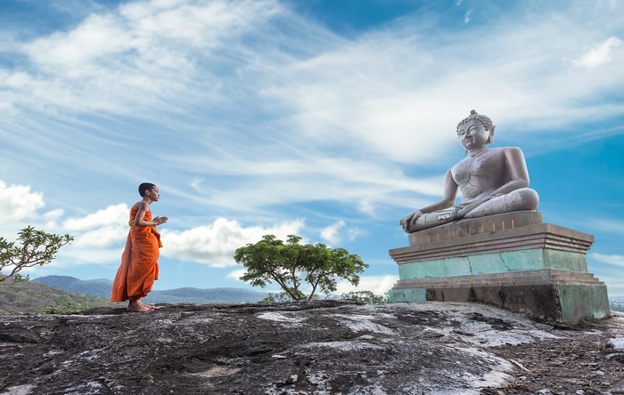Monk praying before Buddha statue on a rocky outcrop
