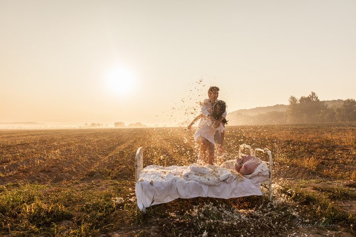 Couple having a pillow fight on a bed in a field at sunset