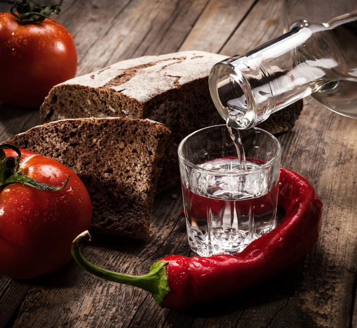 Vodka being poured into a shot glass with bread, tomatoes, and chili pepper