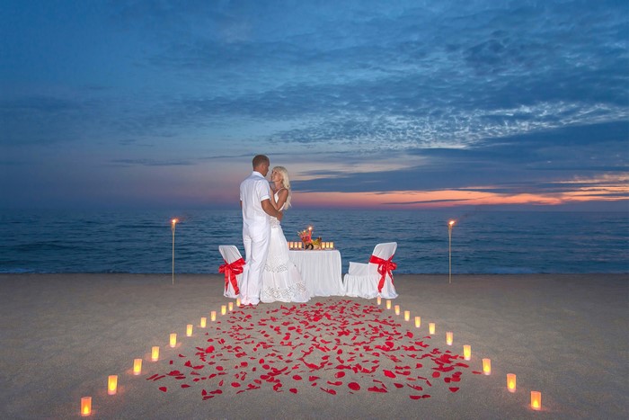 Couple embracing on a beach at sunset with candles and rose petals