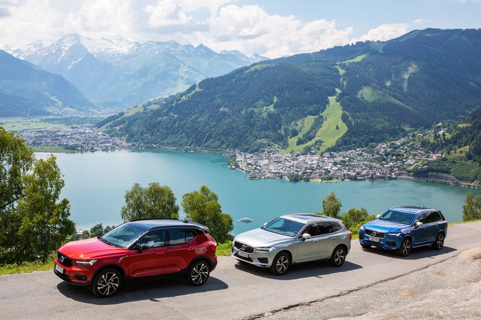 Three Volvo SUVs parked on a scenic mountain road overlooking a lake and town