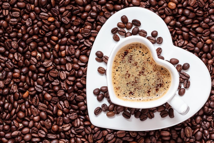 Heart-shaped cup of coffee on a saucer surrounded by coffee beans
