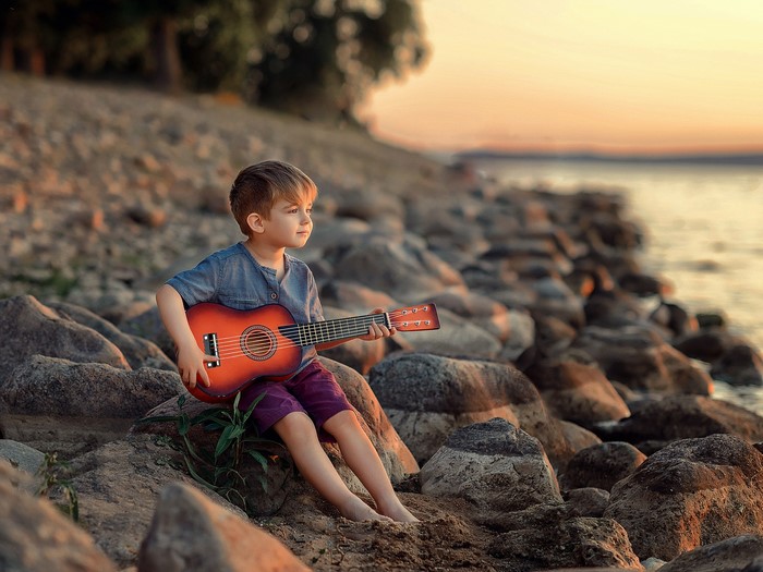 Boy with Guitar on Rocky Shore at Sunset