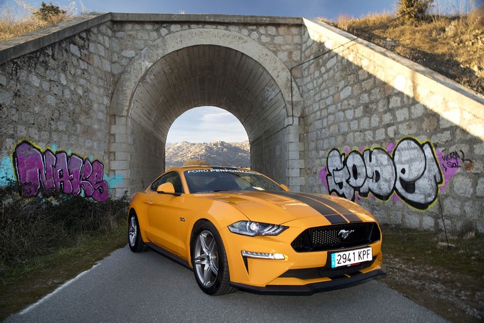 Yellow Ford Mustang GT with racing stripes under a bridge