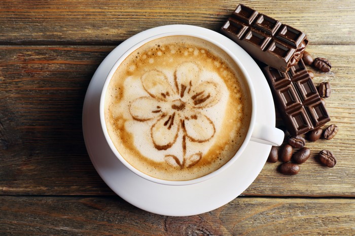 Cappuccino with flower art, chocolate, and coffee beans on wood