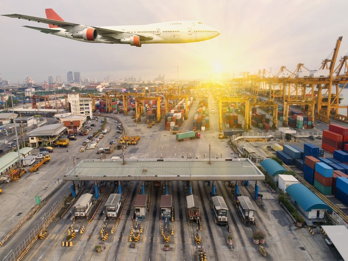 Airplane flying over a busy shipping port with cranes and containers