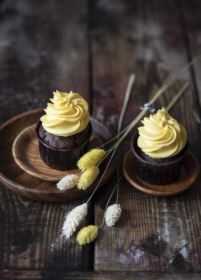 Two chocolate cupcakes with yellow frosting on wooden planks