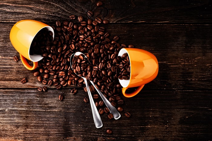 Two orange cups spilling coffee beans with spoons on wood