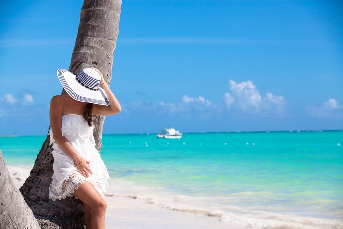Woman in white dress and hat by palm tree on beach