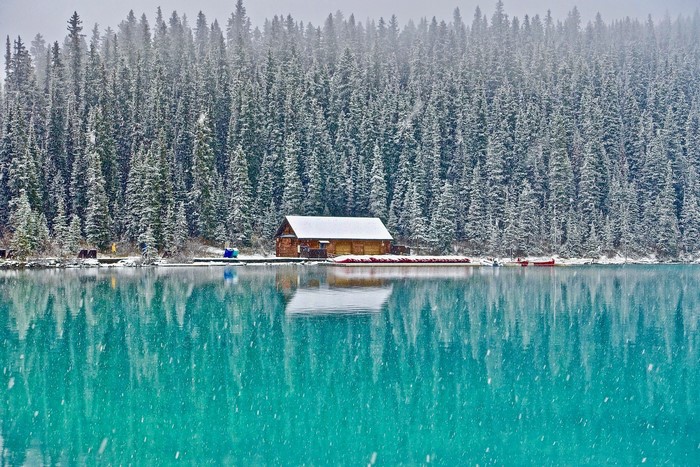 Wooden cabin by a turquoise lake with snow-covered pine forest