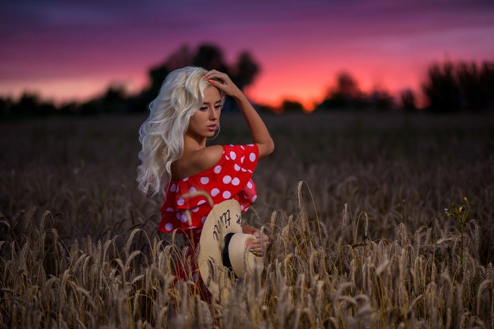 Blonde girl in red polka dot dress holding a hat in a field at sunset