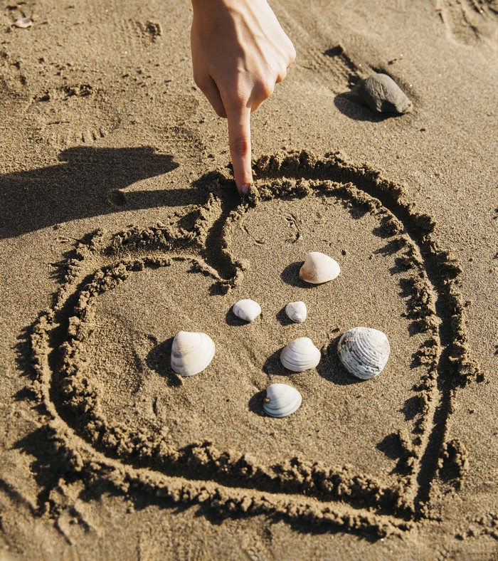 Finger drawing heart in sand with shells