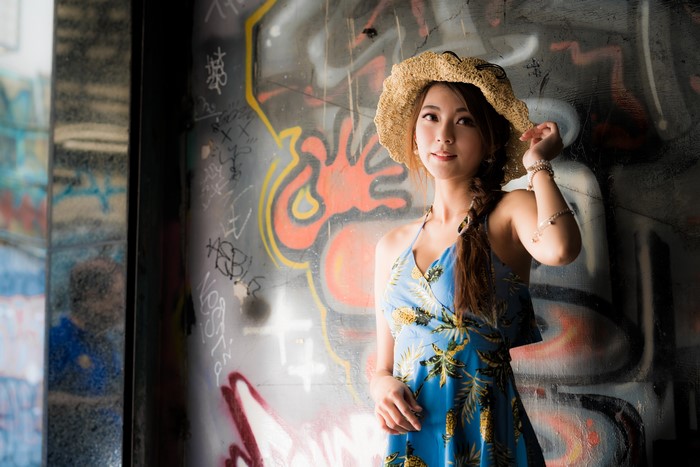 Asian woman with braid hair wearing a hat and dress in front of graffiti wall