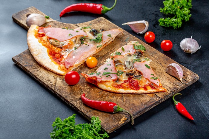 Two slices of ham and mushroom pizza on a cutting board with chili peppers and garlic