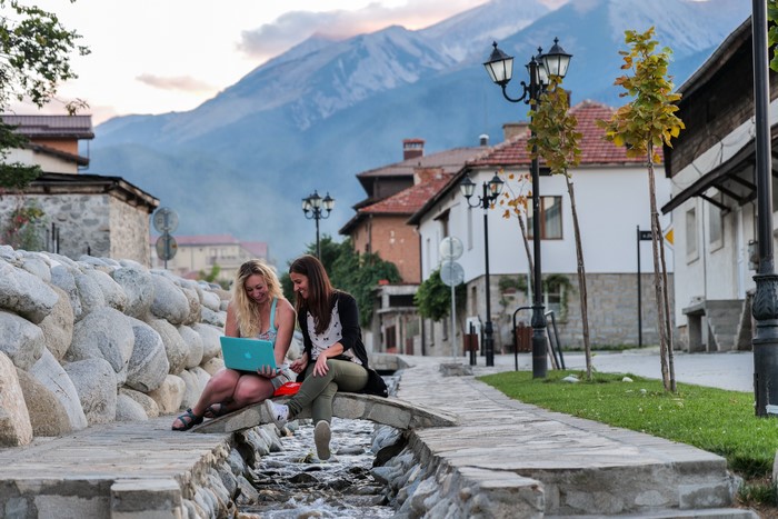 Two girls with laptops sitting on a bridge over a stream