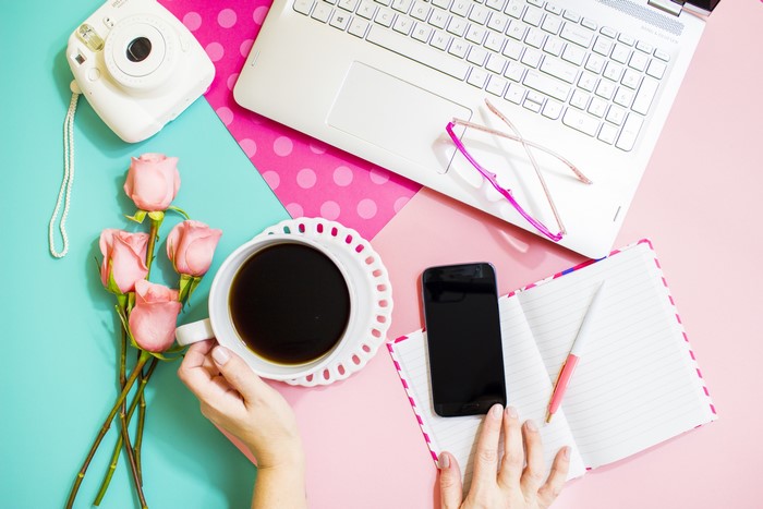 Flat lay of a desk with laptop, camera, coffee, roses, and smartphone