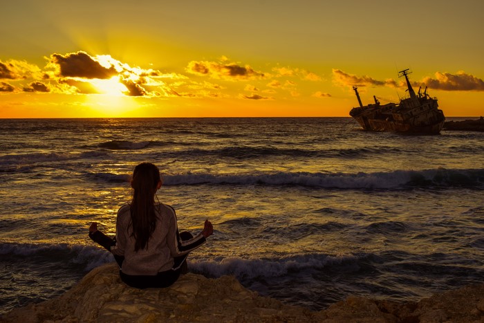 Girl meditating by shipwreck at sunset