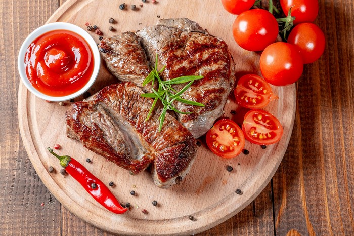 Grilled steaks with tomatoes, chili pepper, and ketchup on a cutting board