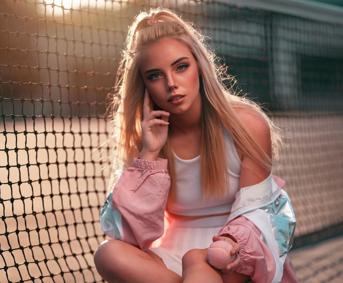 Blonde girl sitting by a tennis net with a ball