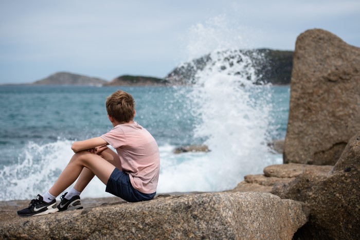 Boy sitting on rocks watching ocean waves crash
