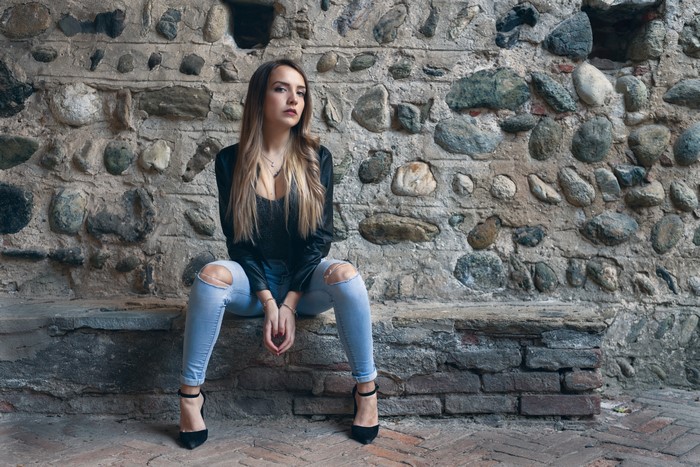 Woman in Jeans and Jacket Sitting by Stone Wall
