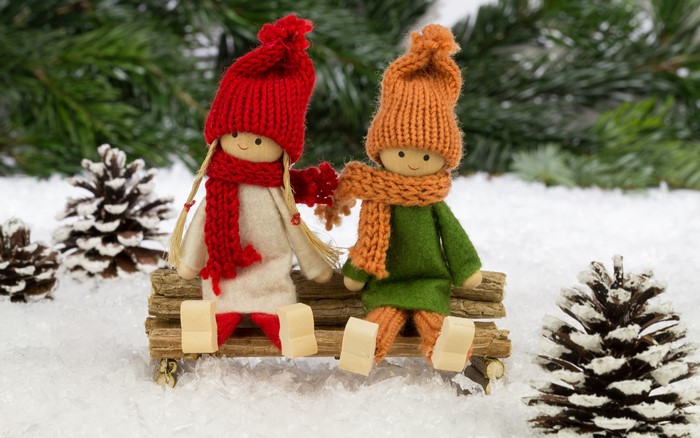 Two dolls sitting on a bench in the snow with pine cones